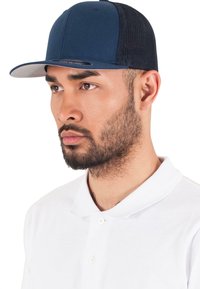 Sötétkék baseballsapka hálós panelekkel, lapos karimával és diszkrét logóval az elülső részen. Fehér galléros inggel viselve.