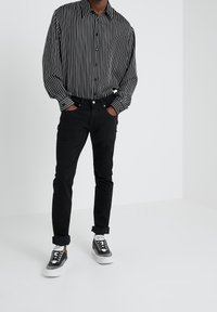 Hombre que lleva una camisa de manga larga a rayas verticales en blanco y negro, pantalones negros con puños y zapatillas en blanco y negro con la marca "BOSS".