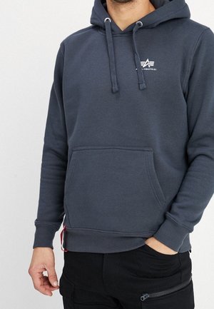 Mann trägt einen dunkelgrauen Hoodie mit Kordelzügen und einer Fronttasche, kombiniert mit schwarzen Hosen, die eine Reißverschlusstasche haben.