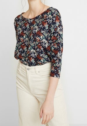 Blumenmuster Langarmtop in Marineblau mit roten, grünen und weißen Mustern; kombiniert mit hellbeigen Cordhose in hoher Taille.