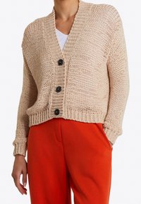 Béžový pletený kardigan s výstřihem do V, zapínáním na knoflíky s černými knoflíky a žebrovanými manžetami, spárovaný s jasně oranžovými kalhotami.