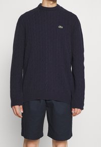 Suéter de punto de cable en color navy con cuello redondo y puños acanalados, que presenta un pequeño logo verde de Lacoste en el pecho. Combinado con shorts oscuros.