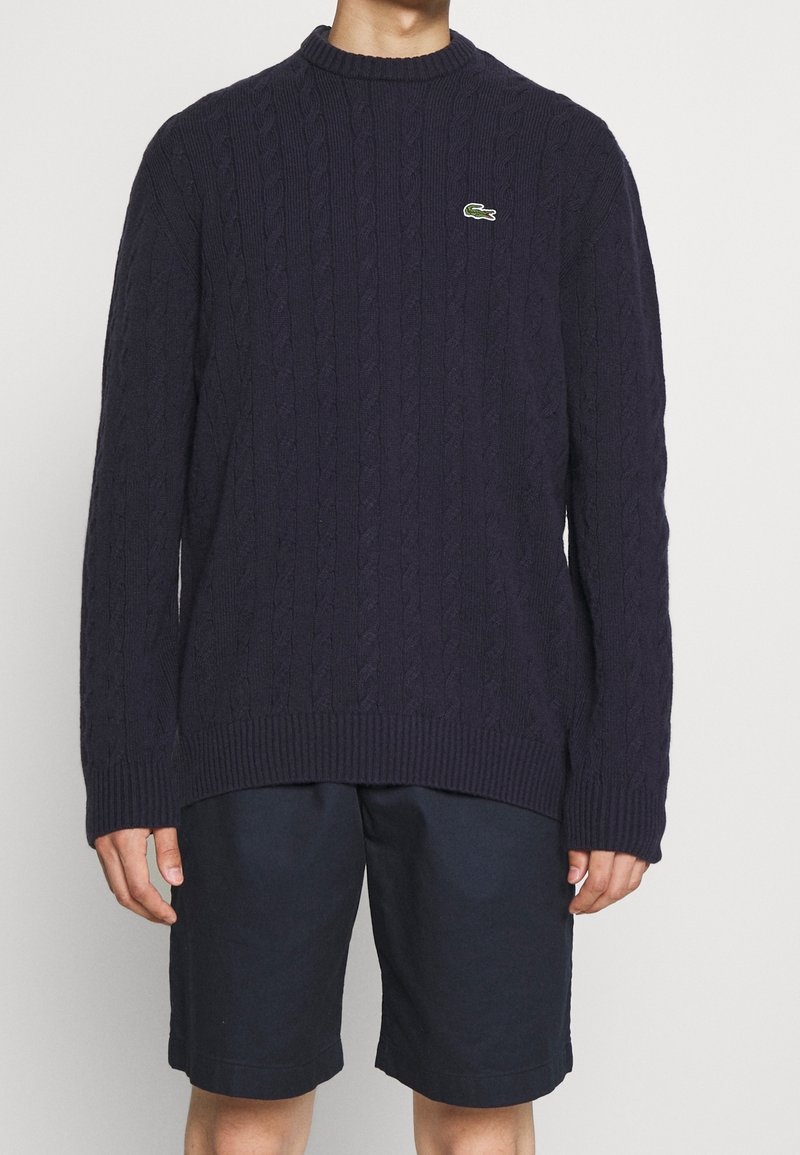 Suéter de punto de cable en color navy con cuello redondo y puños acanalados, que presenta un pequeño logo verde de Lacoste en el pecho. Combinado con shorts oscuros.