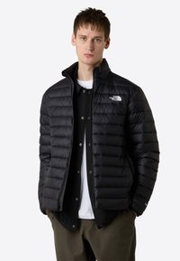 Giacca nera isolata con design trapuntato, colletto alto e chiusura con zip. Presenta un logo sul petto e tasche laterali.