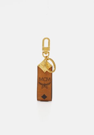 Chaveiro em couro castanho com acabamentos em dourado, apresentando o logo MCM em relevo, um destaque em forma de diamante e um gancho para fácil utilização.
