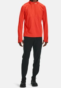 Man som bär en klar orange långärmad sporttröja med kvartsslits, svarta sportbyxor och svarta löparskor.