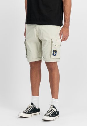 Mann trägt beige Cargo-Shorts mit "Airborne"-Abzeichen auf der rechten Tasche, schwarze Turnschuhe, weiße Socken und ein schwarzes T-Shirt, steht vor weißem Hintergrund.