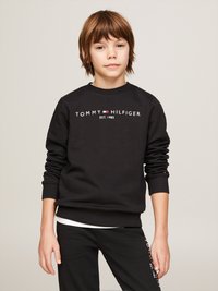 Schwarzer Pullover mit gerippten Bündchen und Saum, der in der Mitte weißen Text "TOMMY HILFIGER EST. 1985" trägt. Über schwarzen Hosen getragen.
