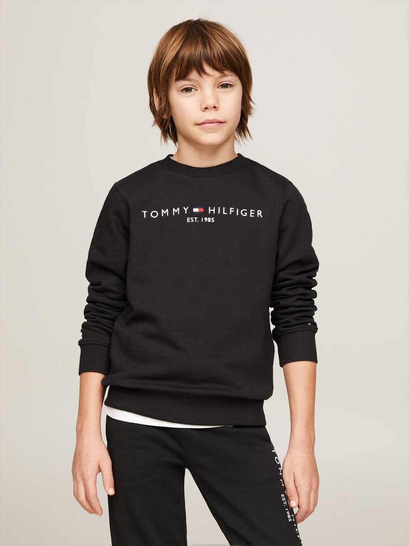 Schwarzer Pullover mit gerippten Bündchen und Saum, der in der Mitte weißen Text "TOMMY HILFIGER EST. 1985" trägt. Über schwarzen Hosen getragen.