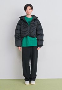 Casaco acolchoado preto curto com gola alta, sobre uma blusa verde de manga curta com o número 23, combinado com calças largas pretas e sapatos brancos.