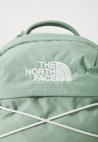 Sac à dos en tissu vert avec un motif en grille, des détails en cordon élastique blanc, et un logo brodé de The North Face.
