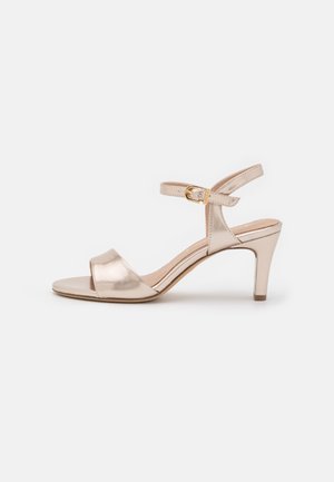 Beige sandal med mellemhøj hæl, åben tå, ankelrem og guldlås, vist på en lys baggrund.