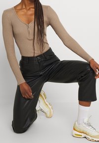 Bruine ribbeltrui met lange mouwen en ritssluiting, gecombineerd met zwarte leren cropped broek en witte sneakers met gele accenten.