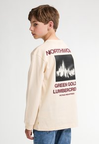 T-shirt a maniche lunghe crema con stampa grafica in bianco e nero sul retro, con testo rosso che recita "NORTHWOOD GREEN GOLD LUMBERCREW."
