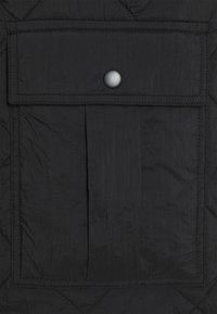 Tessuto nero trapuntato con superficie strutturata, caratterizzato da una tasca frontale chiusa da un bottone a pressione e delimitata da dettagli cuciti.