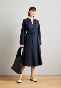Robe rayée marine à manches longues, taille smockée et poches latérales, associée à un sac fourre-tout noir et des ballerines crème avec des accents.
