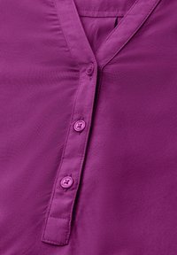 Blusa morada con escote en V, que presenta una fila de seis botones a juego, textura suave y un sutil detalle de fruncido cerca del escote.