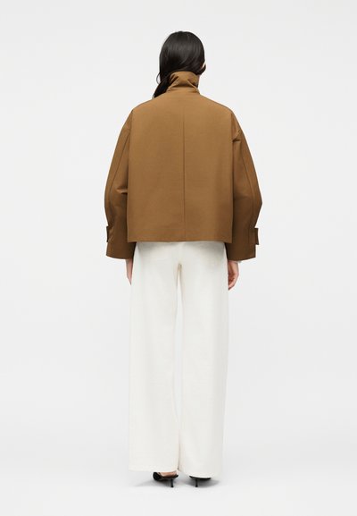 Άτομο με μακριά σκοτεινά μαλλιά που φοράει ένα φαρδύ καφέ μπουφάν, άσπρο παντελόνι με φαρδιές πιέτες και μαύρα τακούνια, στέκεται γυρισμένο προς τα πίσω σε απλό φόντο.