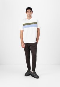 Fehér, rövid ujjú póló, olíva zöld és világoskék vízszintes csíkokkal, sötétbarna, slim-fit nadrággal és textúrázott fekete sportcipővel párosítva.