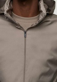 Beige, leichte Zip-Jacke mit hohem Kragen und glatter Textur. Sie verfügt über einen zentralen Reißverschluss und eine mehrlagige Kapuze.