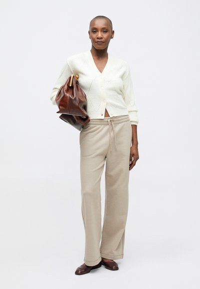 Kremowy sweter z dzianiny w wzór rombów, beżowe spodnie z troczkami oraz brązowa skórzana torebka. Model nosi ciemne buty, stojąc na jednolitym tle.