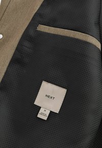 Black and beige jacket interior featuring a textured, honeycomb pattern with a visible pocket and a sewn-in label reading "NEXT," size M.