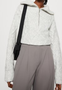 Lichtgrijze gebreide trui met een ritssluiting bij de kraag, voorzien van kabelpatronen en lange mouwen. Gecombineerd met een beige broek met hoge taille.