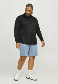 Schwarze, langärmlige Hemdbluse aus glattem Stoff, kombiniert mit hellblauen Denim-Shorts. Modell trägt weiße und schwarze Sneaker mit Socken mit Logo.