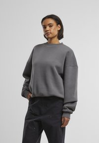 Grå sweatshirt med rund halsudskæring, dropped skuldre og ribbede cuffs. Stoffet ser blødt ud med en glat overflade. Sammen med mørke bukser.