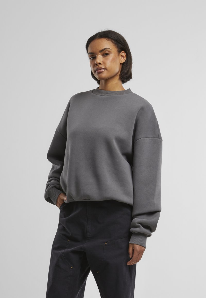 Grå sweatshirt med rund halsudskæring, dropped skuldre og ribbede cuffs. Stoffet ser blødt ud med en glat overflade. Sammen med mørke bukser.