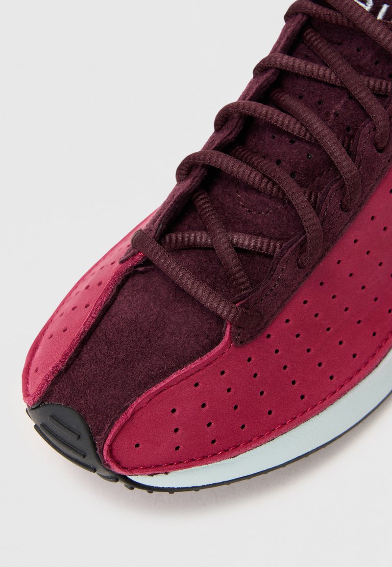 Zapatilla deportiva de color marrón, hecha de materiales de ante y malla, con detalles perforados, cordones planos y suela de goma negra.