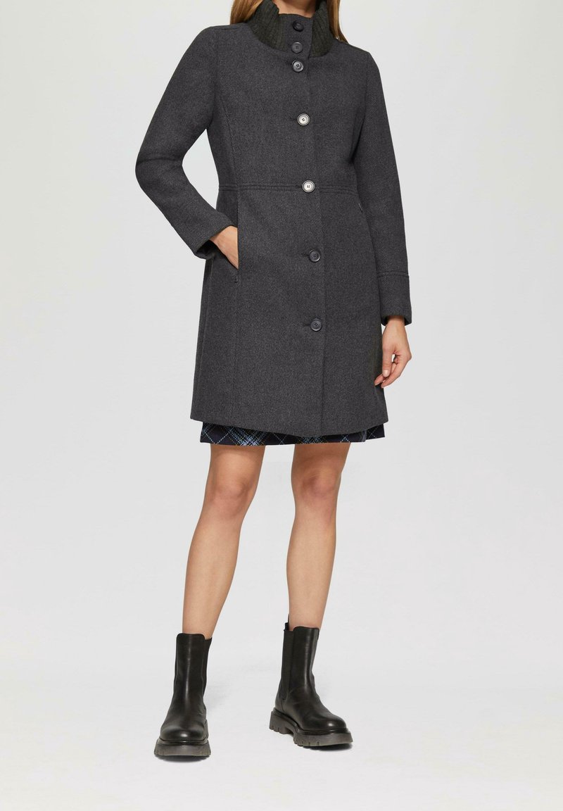 Femme portant un manteau gris foncé boutonné avec un col montant, des bottines noires et une jupe courte à carreaux visible en dessous.
