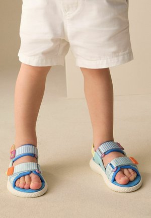 Mažas vaikas dėvi baltus šortus ir mėlynas dryžuotas basutes su oranžiniais ir violetiniais akcentais, stovintis viduje ant kreminės grindų dangos.