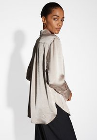 Camicia lunga in satin beige chiaro con una finitura texturizzata, colletto a punta e polsini con bottoni. Orlo arrotondato e vestibilità ampia.