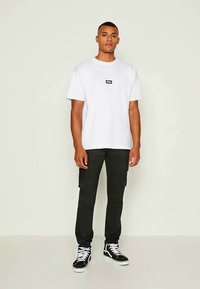 Jovem em pé, vestindo uma t-shirt branca da Obey, calças pretas cargo e ténis de cano alto pretos com atacadores brancos, contra um fundo simples.