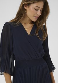 Marineblaues plissiertes Kleid mit transparenten, fließenden Ärmeln, V-Ausschnitt und gerafftem Taillenbund, betont durch filigranen Goldschmuck.