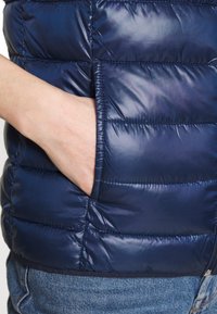 Marineblauer Puffer-Weste aus glänzendem Material mit gesteppten Segmenten. Verfügt über eine Seitentasche und einen Reißverschluss. Leichter Stoff und schlanker Schnitt.