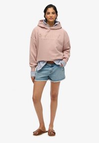Felpa rosa con cappuccio con logo "SuperDry", indossata sopra una camicia a righe blu, abbinata a pantaloni corti di jeans sbiaditi e sandali marroni.