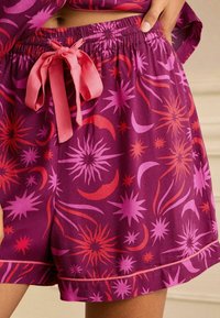 Pantaloni corti viola con un vivace motivo di stelle e lune crescenti, caratterizzati da un fiocco in satin rosa in vita e bordi piped.