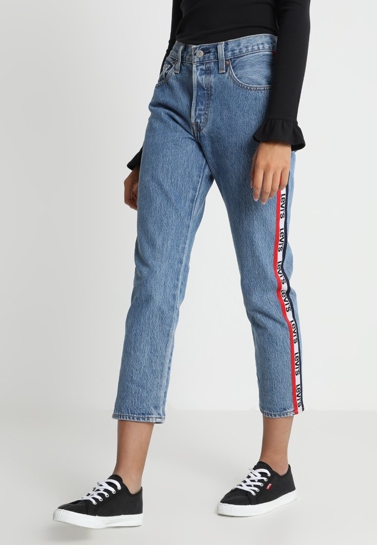 Femme portant un jean court bleu avec une bande aux couleurs rouge et blanche du logo Levi's sur les côtés, associée à des baskets noires et un haut à manches longues noir.
