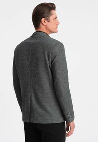 Blazer gris con una tela texturizada, solapa de muesca y ligeras rayas verticales. Presenta puños abotonados y una abertura trasera para mayor movilidad.
