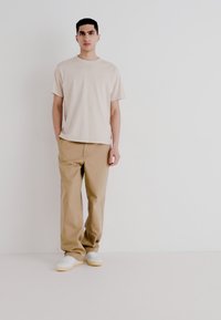 Beige Baumwoll-T-Shirt, lockere Passform; hellbraune Baumwollhose; weiße Sneaker mit gelben Sohlen; glatte Textur, einfaches Design.
