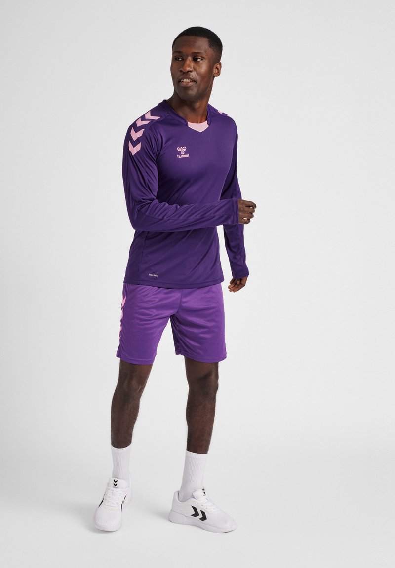 Lilla sport langærmet top med pink chevron detaljer og matchende lilla shorts. Lavet af fleksibelt materiale, parret med hvide atletiske sko.