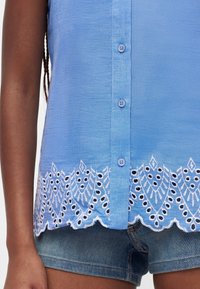 Haut sans manches bleu à boutons avec un ourlet en dentelle blanche brodée, porté avec un short en denim sur une personne à la peau foncée.