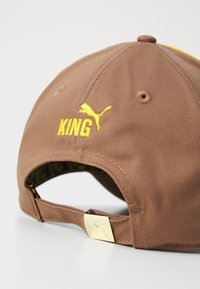Brown baseball cap featuring yellow "KING" embroidery, a Puma logo, and a gold hardware adjuster. Interior has a patterned lining.