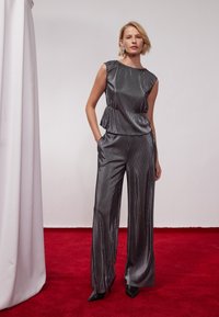 Jumpsuit grigio metallizzato e plissettato con vita a peplum. Presenta pantaloni a gamba larga e design senza maniche. Tacchi neri a punta completano il look.