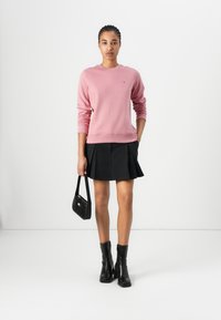 Pink sweatshirt with ribbed cuffs and neckline, paired with a black pleated skirt. Model holds a small black handbag and wears black ankle boots.