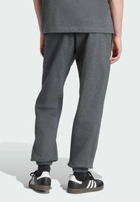 Mörkgrå joggingbyxor i mjukt material, med elastiska muddar, fickor och en liten grön logotyp på högra sidan. Svarta och vita sneakers nedanför.