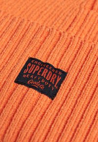 Berretto a costine arancione con etichetta nera. L'etichetta presenta un testo rosso con la scritta "SUPERDRY" e "HEAVY DUTY" in lettere grasse.