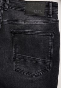 Vaqueros de mezclilla negros con un bolsillo trasero, un acabado texturizado sutil y una etiqueta de cuero con texto en relieve en la cinturilla.